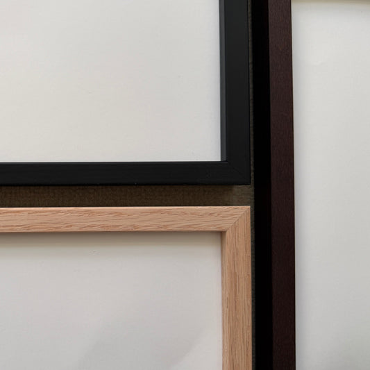Two empty picture frames displayed side-by-side against a white wall, one with a black frame and one with a light oak wood frame.