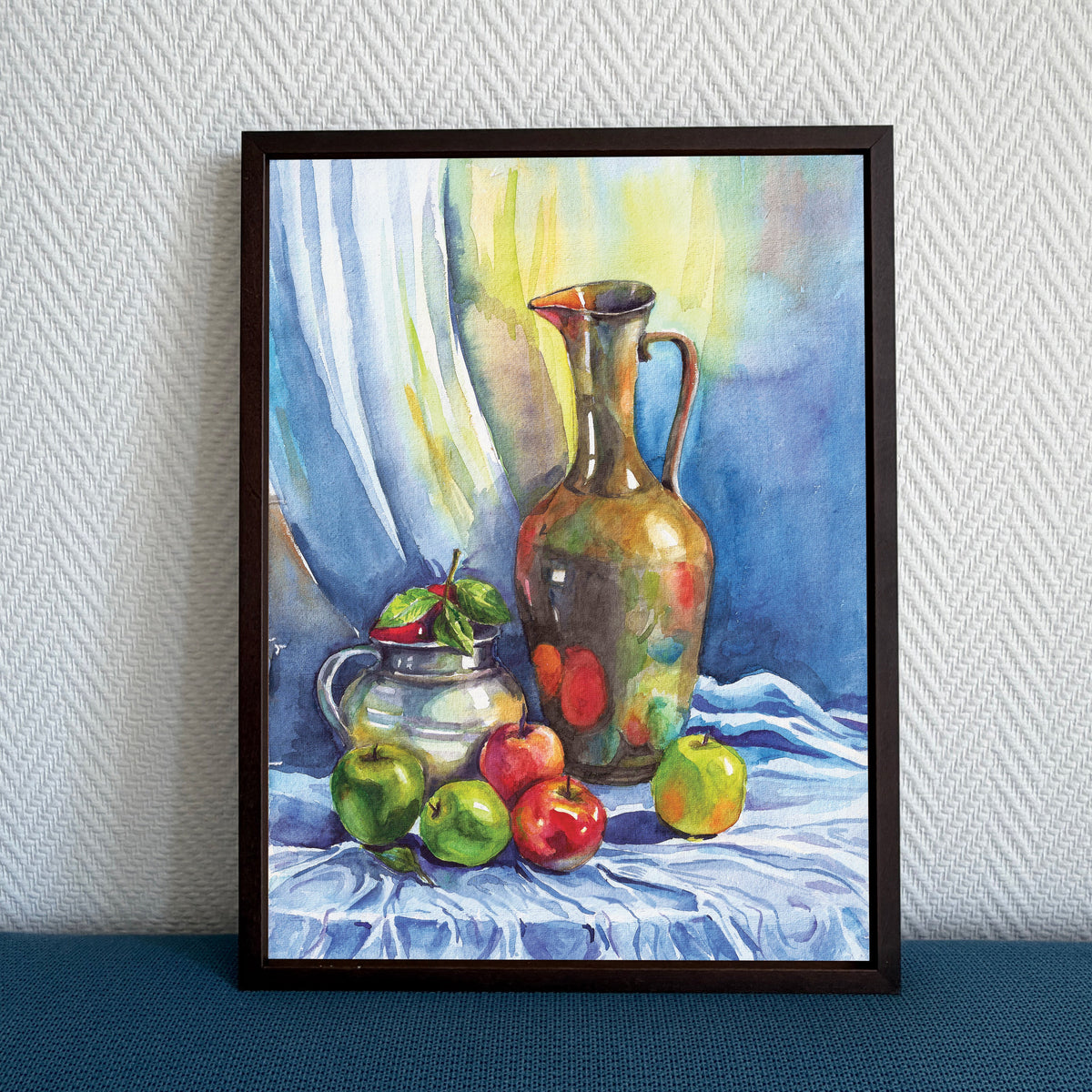 A framed still life painting featuring a patterned brown pitcher, green and red apples, and a small silver pot on a draped table, leaning against a textured light gray wall on a dark blue surface.
